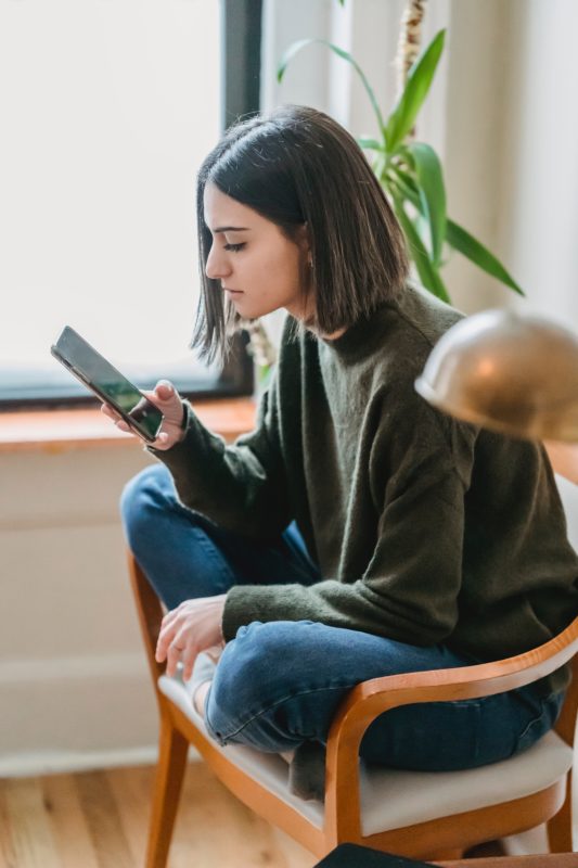 Image of a young woman sitting on a chair looking confused as she checks her phone